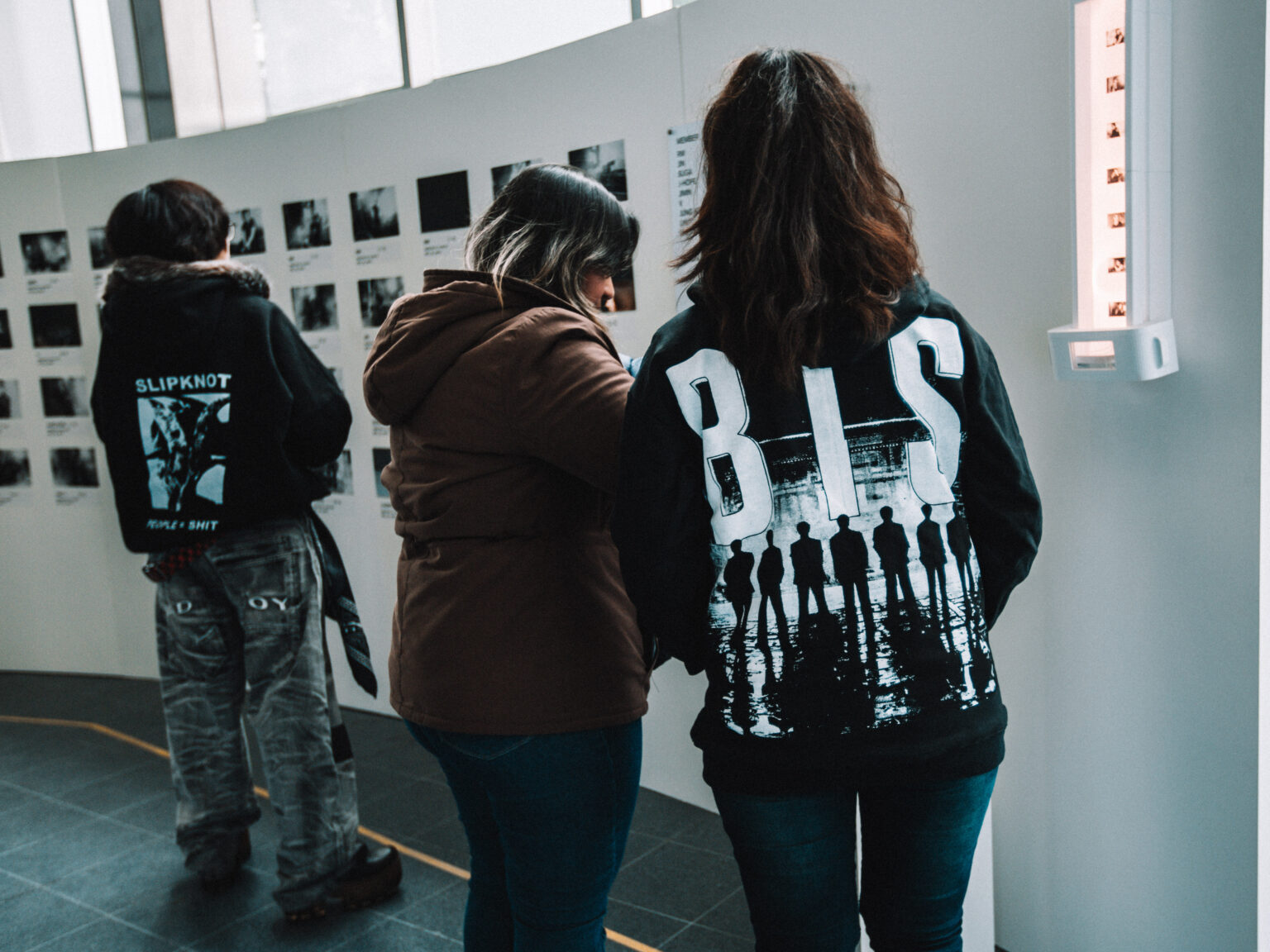 Two fans in BTS jackets looking at the photo wall at the BTS POP-UP: ARIRANG IN BUENOS AIRES, Centro de Convenciones Dr. Arturo Frondizi, Vicente Lopez, 2026 — photographed by Anita Almaraz for K-HipHop Argentina