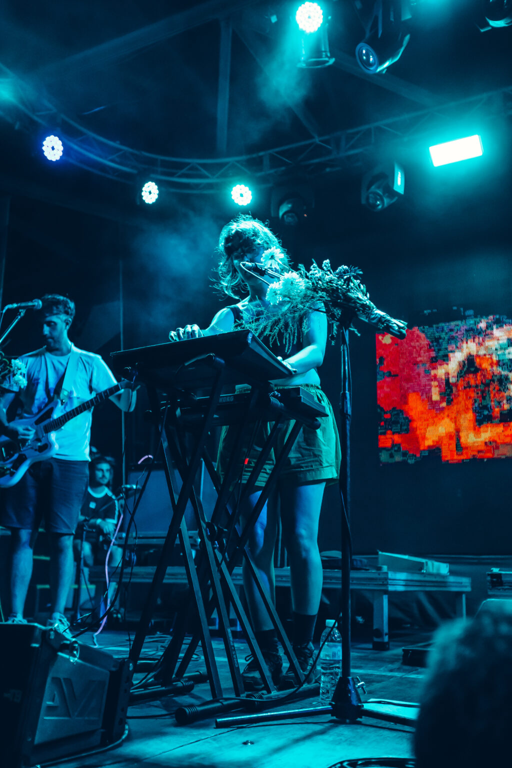 Wide view of the stage, the band playing with flowers tied to the microphone stands and a video screen burning orange behind them &mdash; FestiLaptra at Ciudad Cultural Konex, Buenos Aires, 17 February 2024. Photographed by Anita Almaraz.