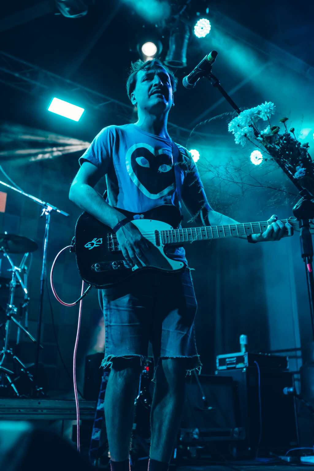 A guitarist looking up with the neck of his guitar raised, flowers on the stand beside him &mdash; FestiLaptra at Ciudad Cultural Konex, Buenos Aires, 17 February 2024. Photographed by Anita Almaraz.