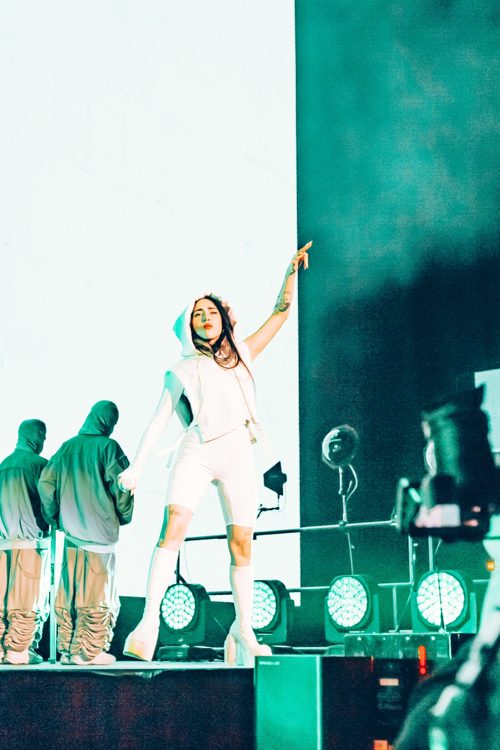Nicki Nicole raising her arm to the crowd under teal stage lighting — Movistar Arena, Buenos Aires, August 2023. Photographed by Anita Almaraz for Agencia Télam.