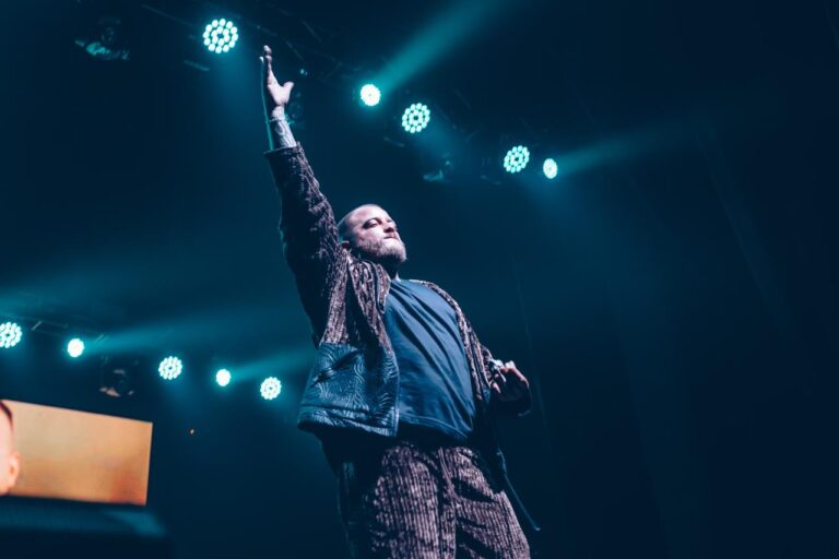 The artist with an arm raised against the blue beams — Zeballos presenting Asimetría at Vorterix, Buenos Aires, 2026. Photographed by Anita Almaraz.